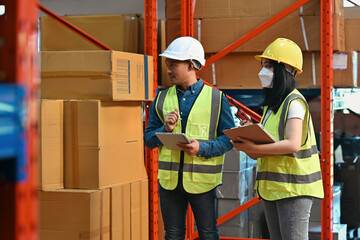 Two warehouse employees wearing safety gear and helmets, holding clipboards and a walkie-talkie,...