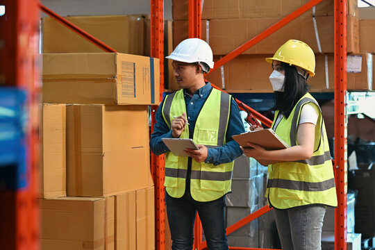 Two warehouse employees wearing safety gear and helmets, holding clipboards and a walkie-talkie, planning logistics inside a storage facility