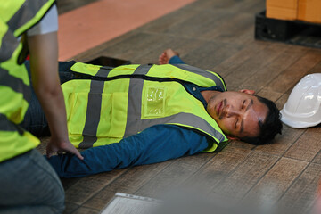 A worker lies unconscious on the floor of a warehouse while his coworkers check his pulse as part of a first aid procedure