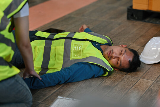 A worker lies unconscious on the floor of a warehouse while his coworkers check his pulse as part of a first aid procedure