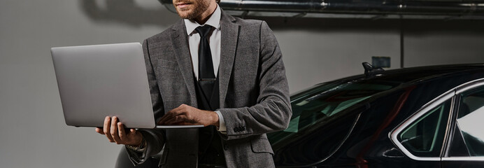 A well-dressed businessman with a beard is focused on his laptop near his luxury car, banner