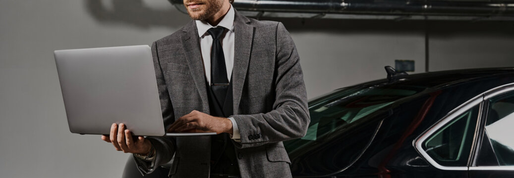 A well-dressed businessman with a beard is focused on his laptop near his luxury car, banner