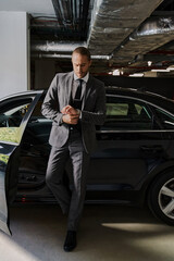 A well-dressed businessman with a beard checks his watch by his luxury car in a garage.