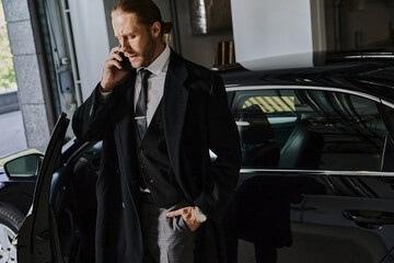 A handsome businessman dressed in an elegant suit stands by his car, talking on the phone.