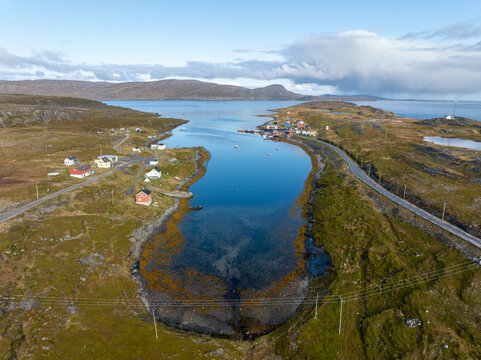 Aerial view of a serene fjord reflecting the sky, dotted with colorful houses and connected by a winding road, Gamvik, Finnmark, Norway.