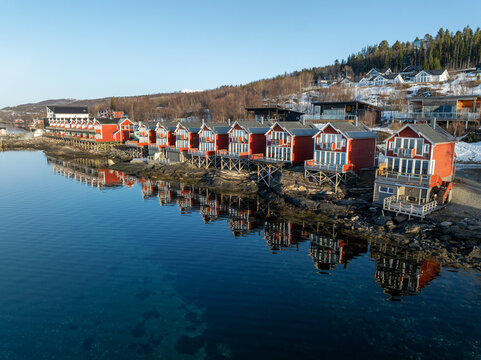 Aerial view of vibrant red cabins with white trim mirrored in the serene, deep blue waters of the Norwegian fjord, Troms&Atilde;&cedil;, Norway.