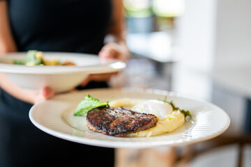 Close-up of a person holding two plates with gourmet meals, featuring steak, mashed potatoes, poached egg, and garnish, served in a restaurant setting.