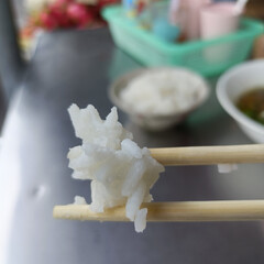 Close up of piece of white fish being held with chopsticks, with bowls of rice and soup background, table casual dining setting, capturing moment of eating traditional Asian cuisine, with focus