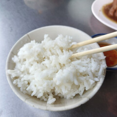 Bowl of fluffy white rice with chopsticks resting on top, served on gray table with other dishes partially visible in background, evoking simple and comforting meal experience