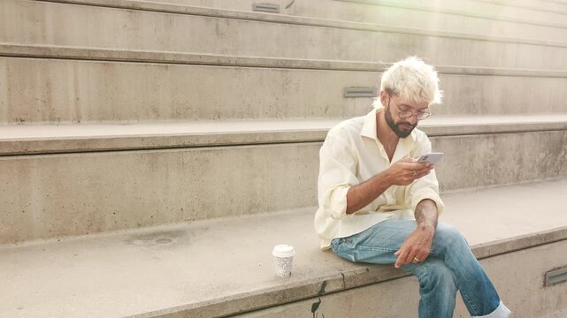 A young man with bleached blond hair sits on concrete steps, enjoying a coffee and scrolling through his phone. He smiles while reading a text or social media post.