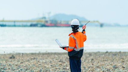 Female environmental engineer, ecological scientist industrial worker working on laptop and talking on radio inspect water quality at industry construction site near oil refinery plant by the sea.