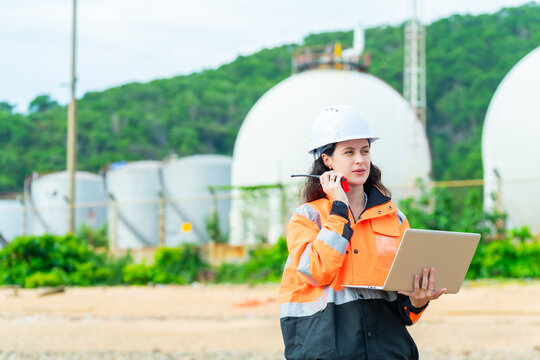 Female environmental engineer, ecological scientist industrial worker working on laptop and talking on radio inspect water quality at industry construction site near oil refinery plant by the sea.