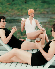Woman in white swimsuit holding martini seated among two men in black vintage outfits. Concept of...