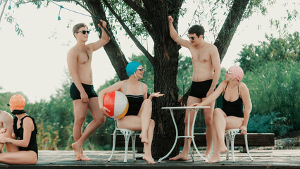Group of young people in vintage swimwear relaxing by lakeside under tree. Concept of lifestyle and...