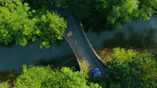 Light blue convertible driving over narrow bridge surrounded by trees and water. Small cabriolet moving across river crossing top down view. Blue open top car traveling between green forest and calm