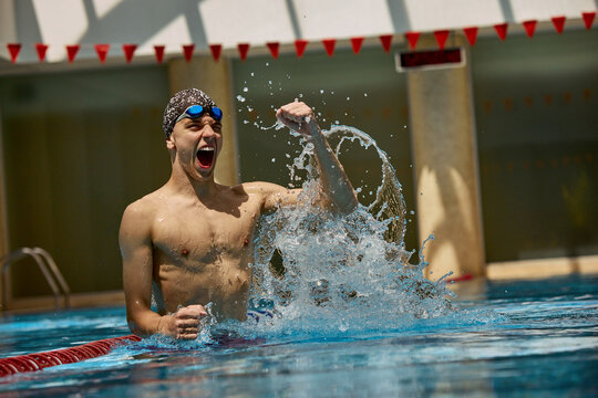 A triumphant swimmer, a young Caucasian male, celebrates victory in an indoor pool with a raised fist and splashing water. Concept of achievement and sportsmanship celebration.