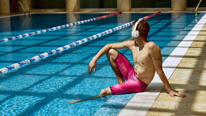 A Caucasian man relaxes poolside at an indoor swimming facility, using headphones and wearing...