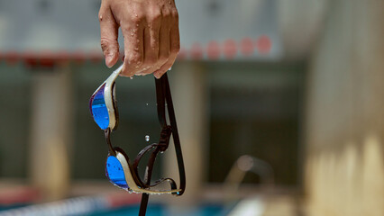 A person holds a wet pair of swimsuit goggles by the pool edge, the background blurred. Concept of...