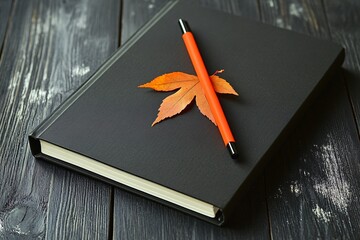 Black notebook with orange pen and autumn leaf on wood