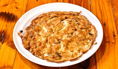 A close-up shot of a golden-brown, fluffy omelette served on a simple white plate. The omelet has a irregular, rustic shape with crispy, darker edges and a soft, airy texture in the center. 