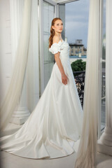 Fototapeta premium Elegant bride in a flowing white gown poses gracefully near an open window with a city view at dusk