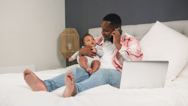 Father Multitasking While Caring for Infant at Home with Smartphone and Laptop on Bed