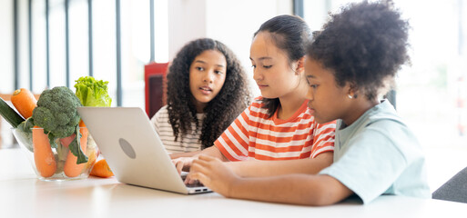 Diverse group of kids three girls happy looking at computer laptop studying nutritional value of fruits in modern workplace learning. Table while sharing ideas in classroom school.