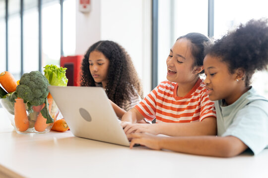 Diverse group of kids three girls happy looking at computer laptop studying nutritional value of fruits in modern workplace learning. Table while sharing ideas in classroom school. - Powered by Adobe