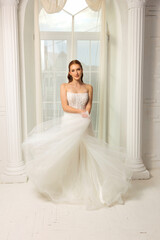 Elegant bride in flowing wedding dress poses near window in bridal studio for a timeless portrait