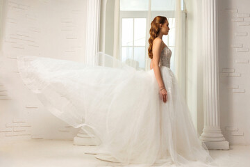 Elegant bride in flowing white gown stands near window with draping fabric in bright, airy setting