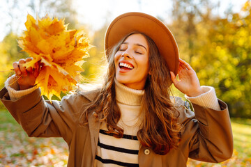 A stylish woman in a hat and holding a bouquet of fallen leaves enjoys the sunset in an autumn park. A beautiful woman has fun and walks outdoors. Seasonality concept. Lifestyle.
