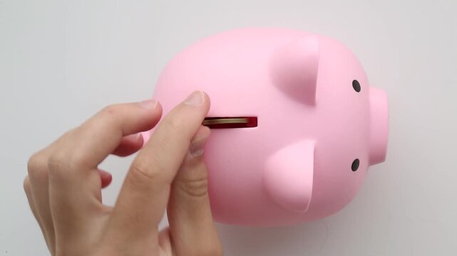 Human hand carefully placing multiple coins into pink ceramic piggy bank slot with audible metallic sounds on neutral background surface