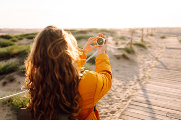 Close-up of a young female traveler's hands holding a compass against the backdrop of a sunset beach. A woman in a bright jacket looks at direction of travel and enjoys sunlight. Travel concept.
