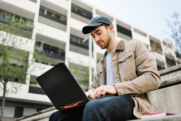 Male student or young freelancer typing on laptop, working remotely outdoors in city