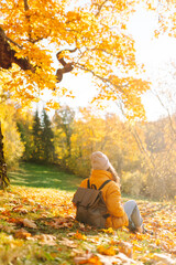 A cheerful woman in a bright jacket collects and throws bright foliage in a sunny park. A beautiful woman has fun and enjoys the autumn landscape. The concept of a walk, travel and relaxation.