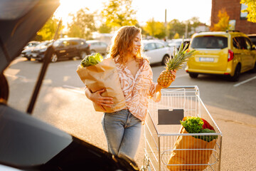 Fototapeta premium Cute woman with bag of fresh vegetables and fruits putting in trunk at sunset. Happy housewife with pineapple in hands and bag full of products in parking lot near car. Shopping, routine concept.