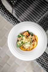 Top view of a white plate with couscous salad, spinach, cherry tomatoes, and roasted yellow squash on a wicker chair, showcasing a healthy and vibrant meal outdoors.