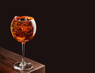 A cocktail with ice served in a large stemmed glass on a dark wooden bar with a black background. copy space for text