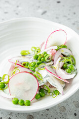 Fresh vegetable salad with radishes, peas, cucumbers, and green onions in creamy dressing, served in a white bowl on a gray textured surface