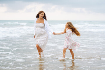 Young pregnant mother with daughter walking on sandy beach. Happy woman with child having fun and enjoying time together. Childhood, pregnancy and nature concept.