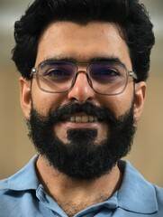 Headshot portrait of a friendly smiling South Asian man with a beard and glasses.