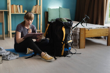 female traveler preparing for hiking trip, packing trekking equipment into large backpack at home,...