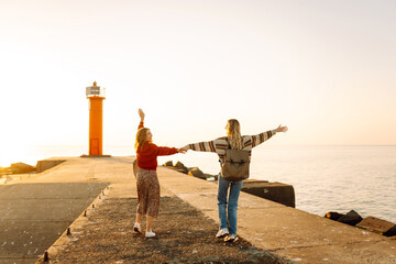 Fototapeta premium Two female friends having fun and walking along the pier against the background of the lighthouse. Beautiful women enjoying the sunset and time together outdoors. Concept of friendship, walks.