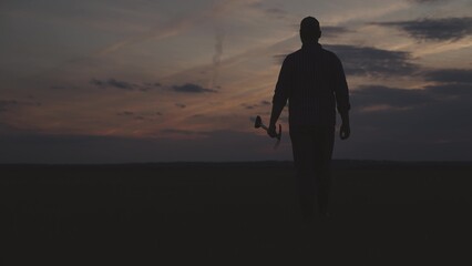 farming, silhouette of man walks with shovel in his hands across field against background of sunset...