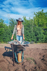 Female farmer in rubber boots carrying wheelbarrow against sky. Organic gardening. Front view