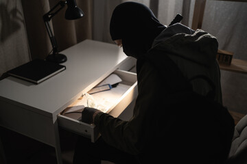 Illegally breaking. Male robber wearing black balaclava steals money from desk at apartment. Thief...