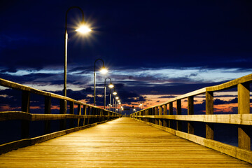 Obraz premium An illuminated pier disappears into the darkness, lanterns illuminating the way under a dramatically cloudy sky. Sun and clouds at dawn on the Baltic Sea beach of 18586 Göhren, Rügen Island