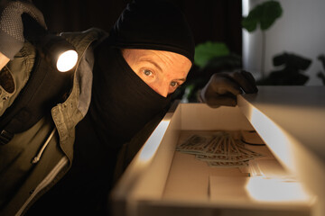 Illegally breaking. Male robber wearing black balaclava steals money from desk at apartment. Thief...