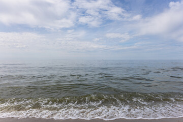 Calm sea water surface with small waves and foam on sandy shoreline under blue cloudy sky.