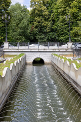Urban canal with fountain jets and stone bridge in city park.